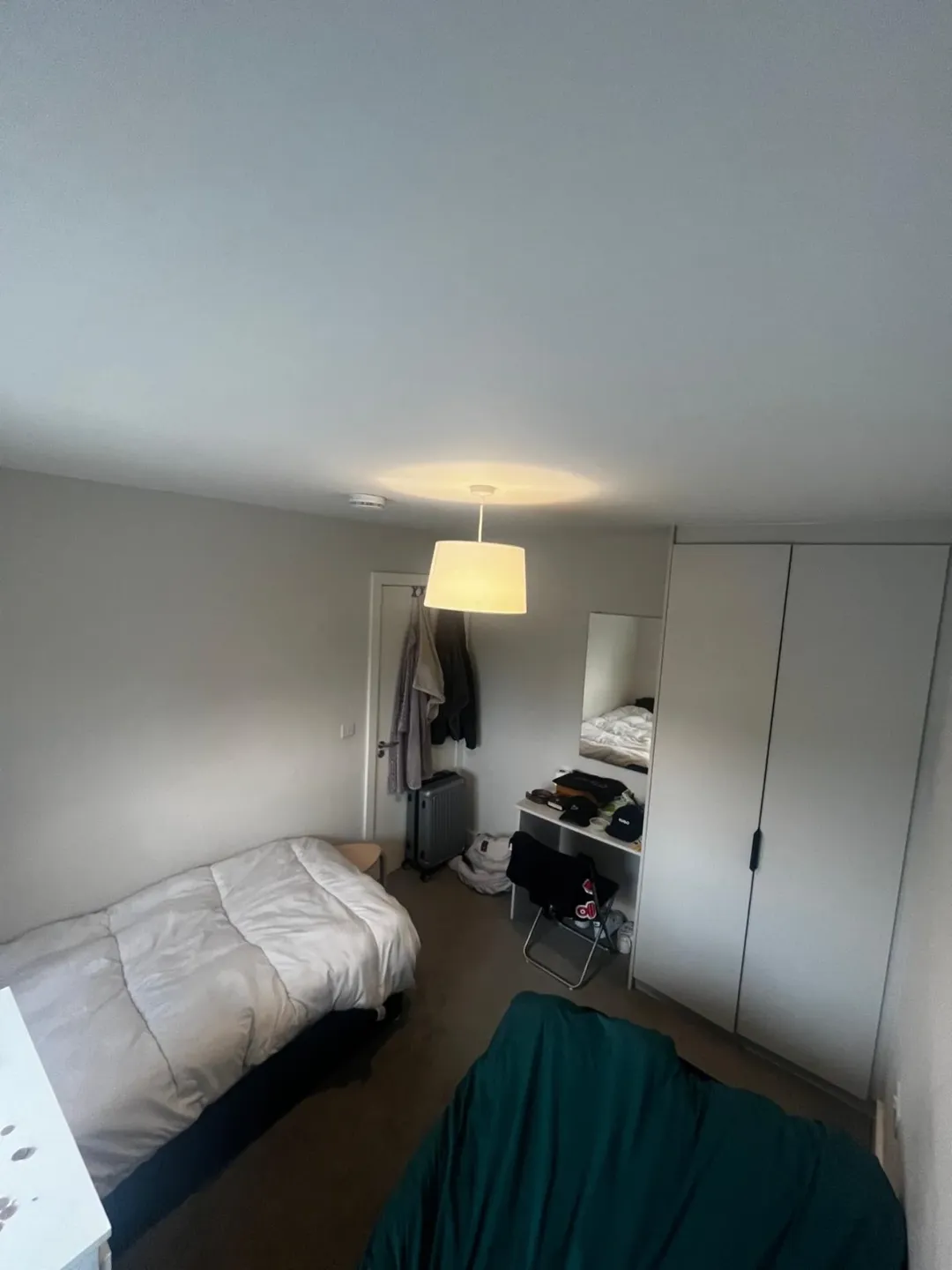 Cozy bedroom with bed, wardrobe, desk, and hanging clothes. Bright overhead light.