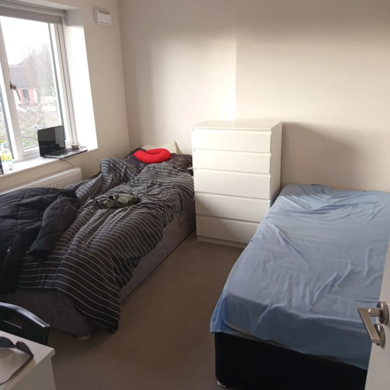 Bedroom interior with two single beds, white dresser, window, and neutral decor.