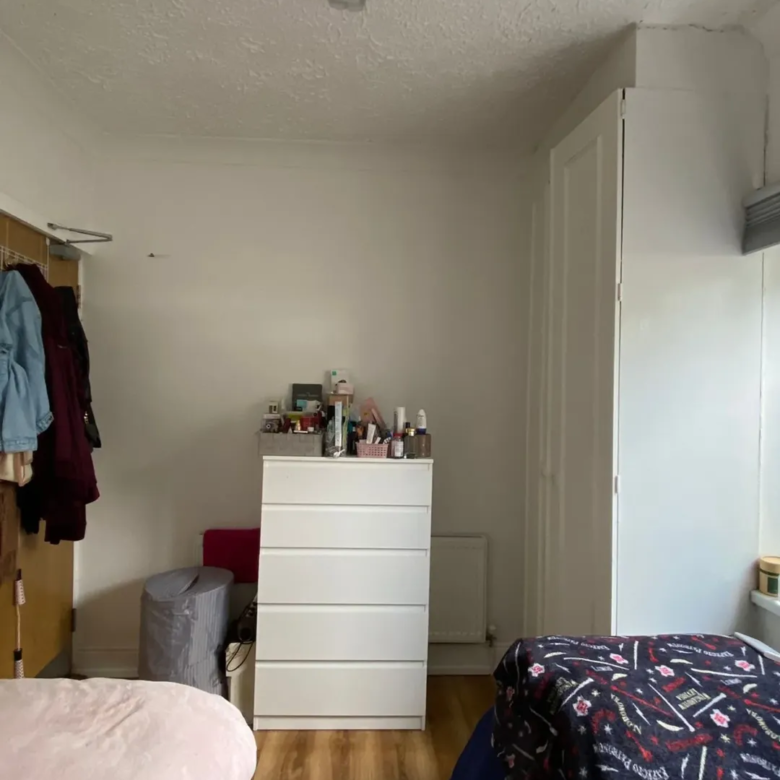Bedroom with white dresser, wardrobe, bed, and clothes rack against a white wall. Natural light from window.