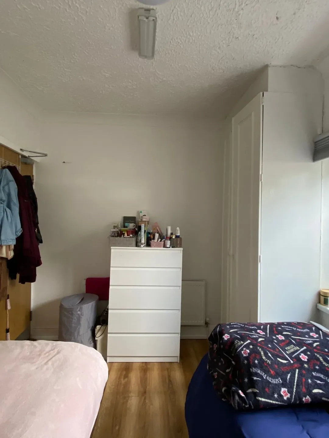 Bedroom with white dresser, wardrobe, bed, and clothes rack against a white wall. Natural light from window.