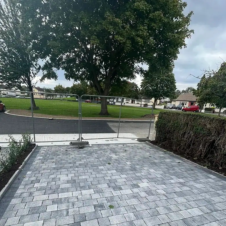 Gray brick driveway with temporary fencing and green trees in the background.