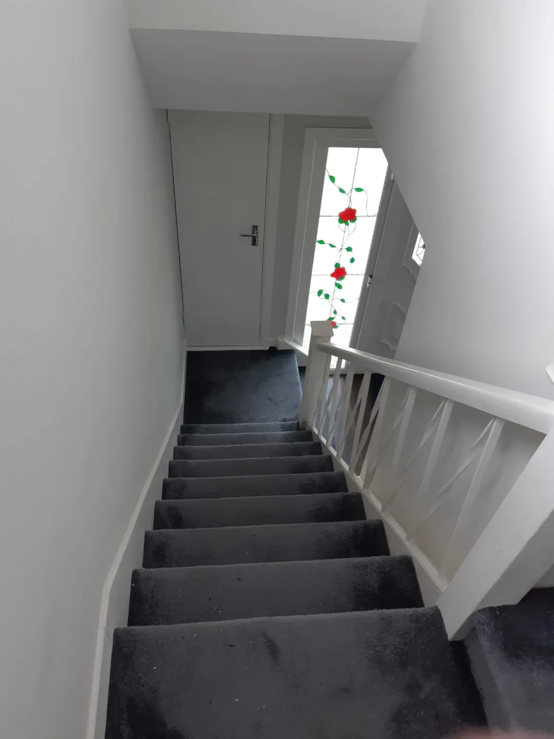 Stairway with gray carpet and white banister leads to a door with rose stained glass.