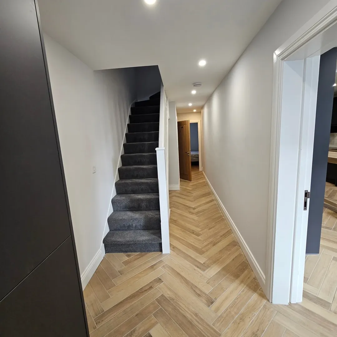 Hallway Herringbone Floor Carpeted Stairs Hallway with herringbone wood floor, carpeted stairs, and white walls.