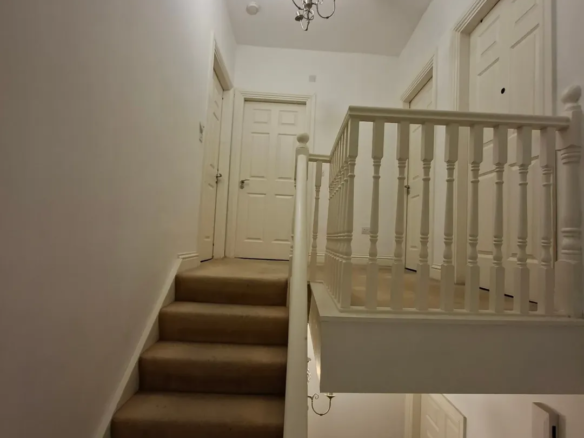 Hallway with staircase, white walls, and neutral carpet. Stairwell with white banister and closed doors.