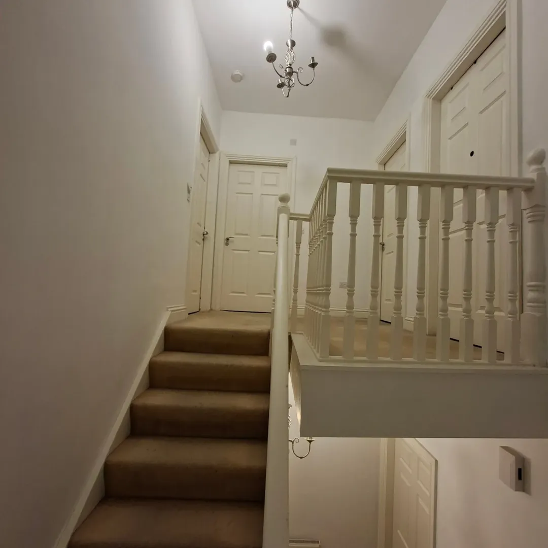 Hallway with staircase, white walls, and neutral carpet. Stairwell with white banister and closed doors.