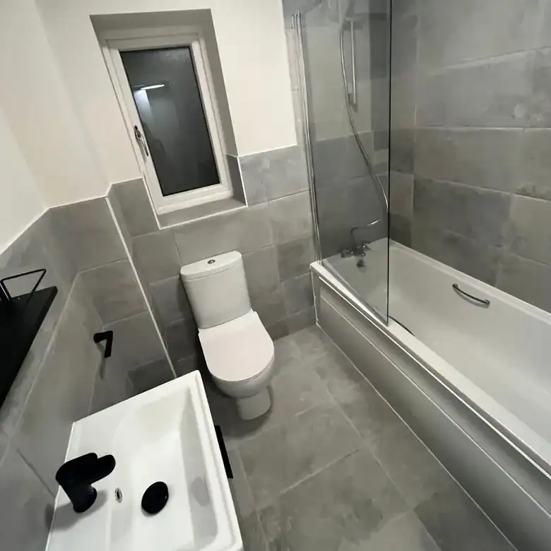 Modern bathroom with gray tile, white toilet, sink, and bathtub with glass screen.