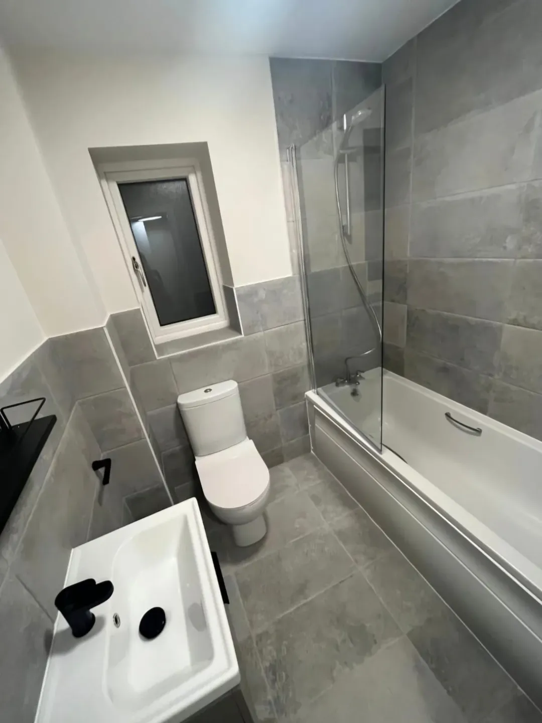 Modern bathroom with gray tile, white toilet, sink, and bathtub with glass screen.