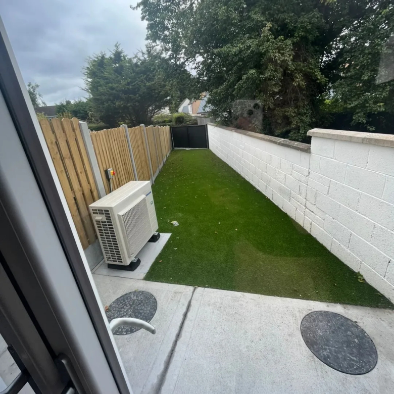 Small backyard with artificial grass, white wall, wooden fence, and air conditioning unit.