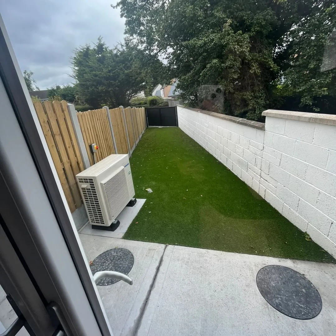 Small backyard with artificial grass, white wall, wooden fence, and air conditioning unit.