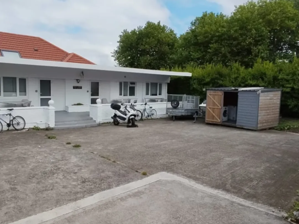 White house with red roof, scooter, bikes, trailer, and shed in driveway under cloudy sky.