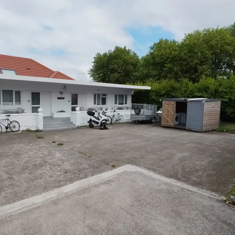 White house with red roof, scooter, bikes, trailer, and shed in driveway under cloudy sky.
