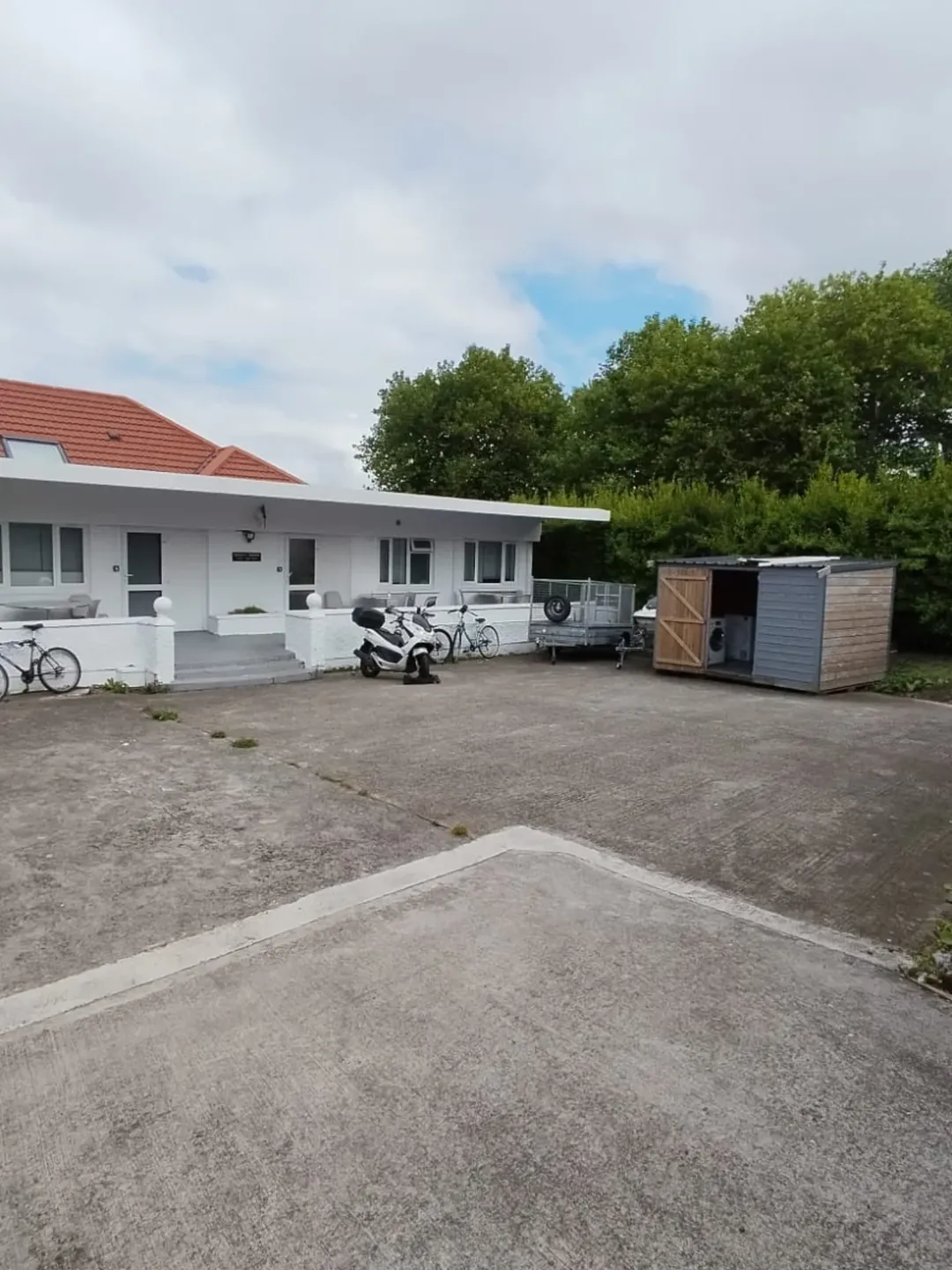 White house with red roof, scooter, bikes, trailer, and shed in driveway under cloudy sky.