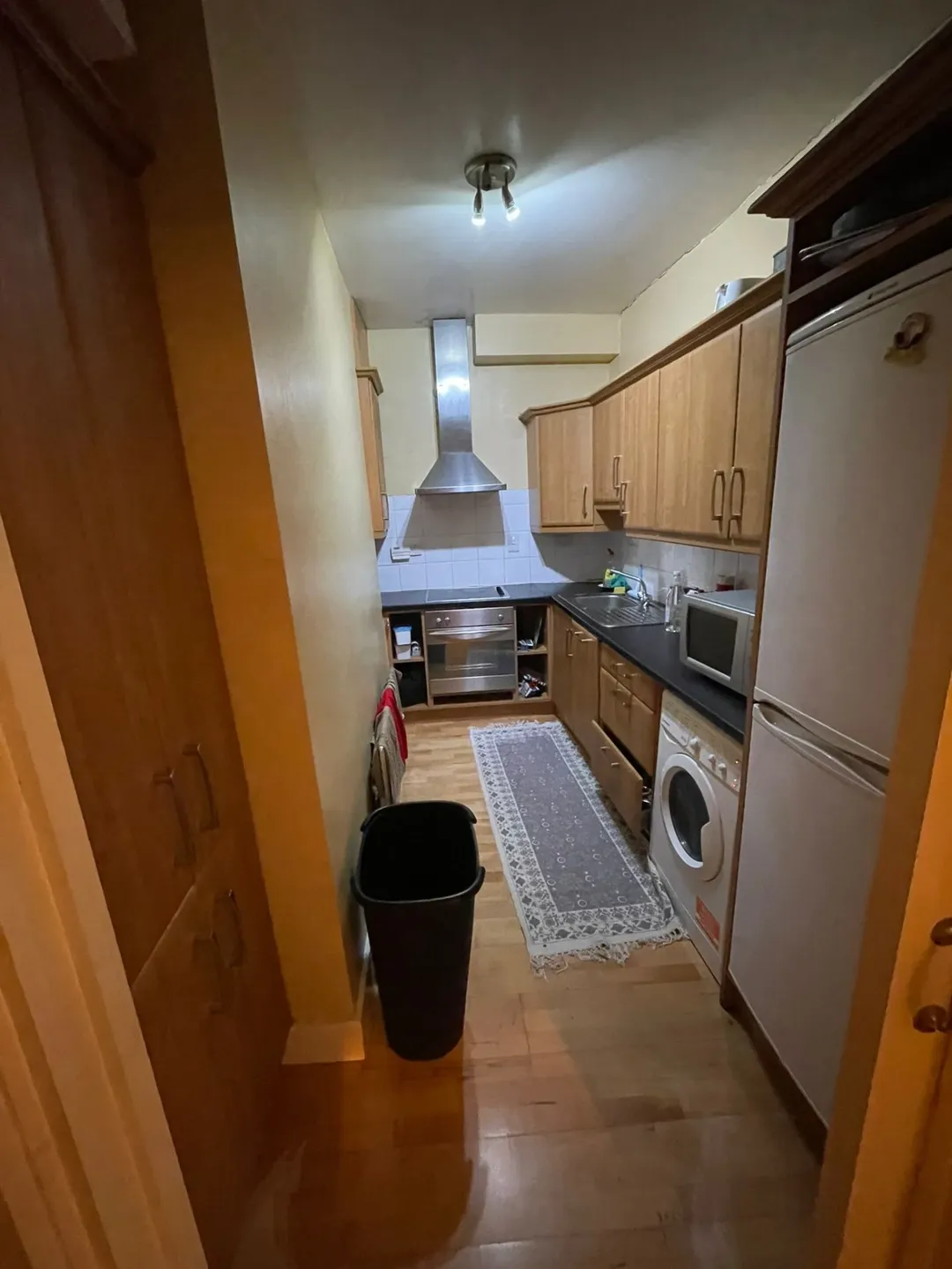Narrow kitchen with wood cabinets, appliances, gray rug, and black trash can.