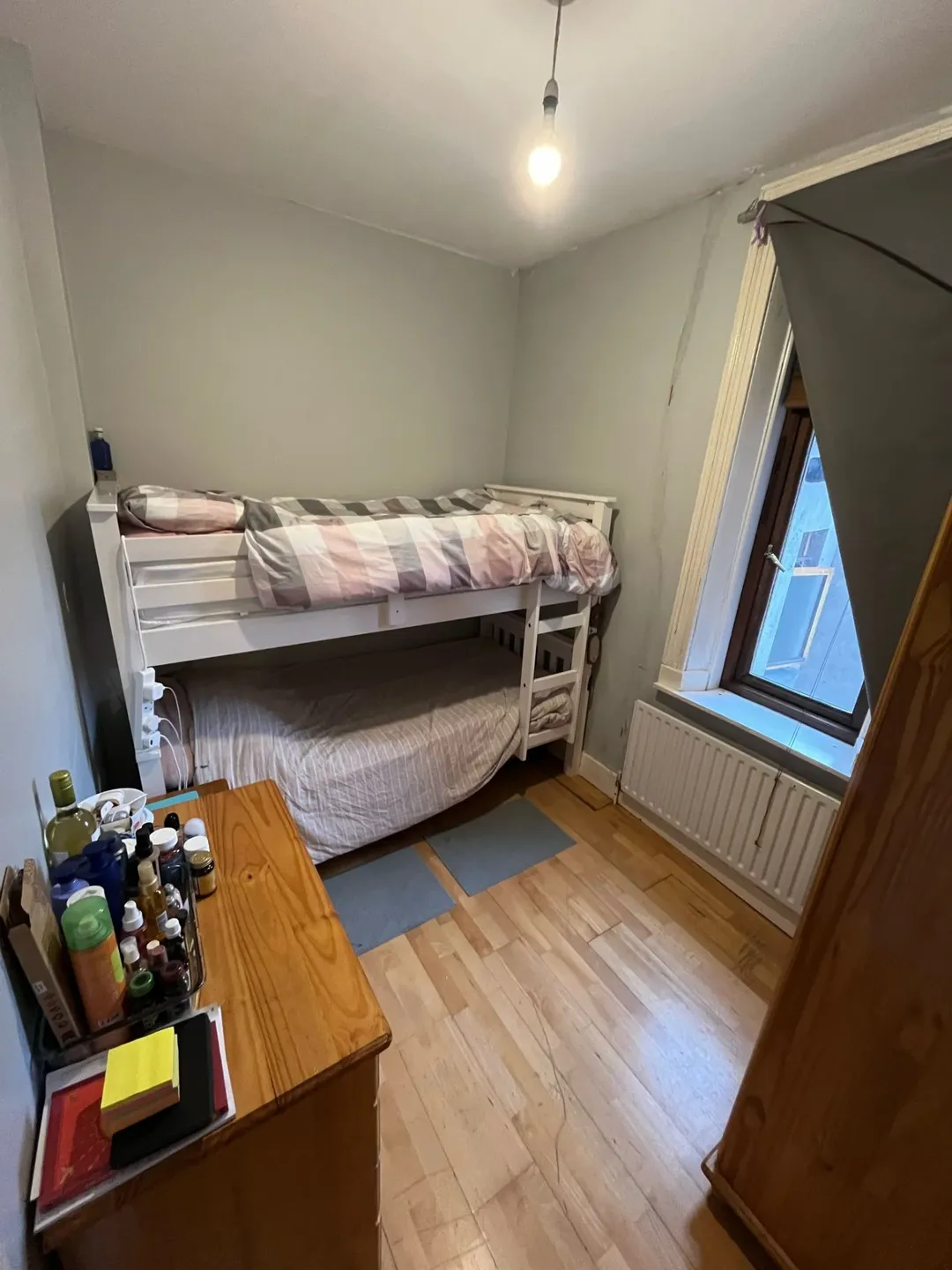 Small bedroom with white bunk bed, wooden floors, and window with grey curtains.