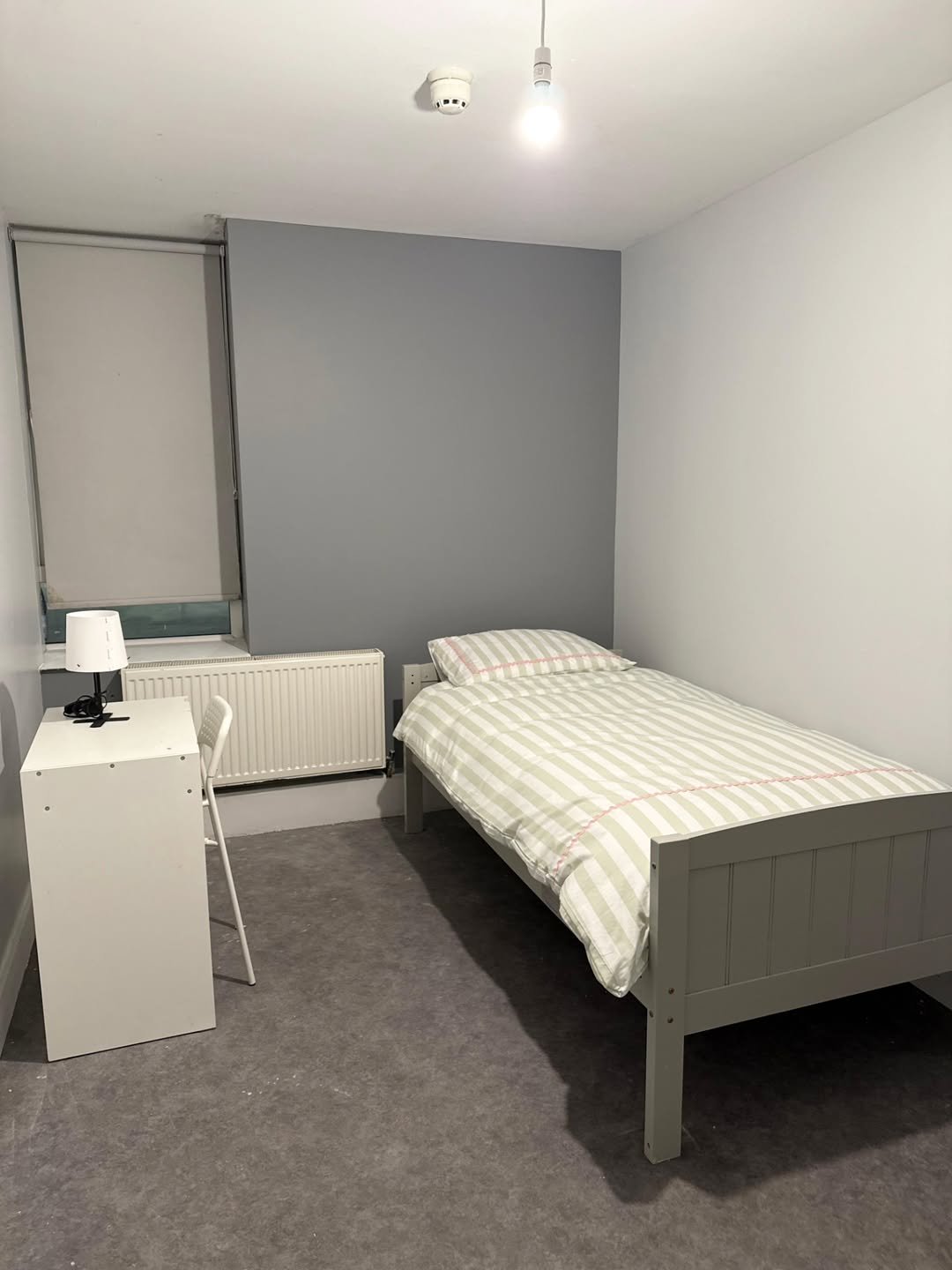 Small bedroom with single bed, desk, radiator, and grey accent wall. Minimalist interior design.