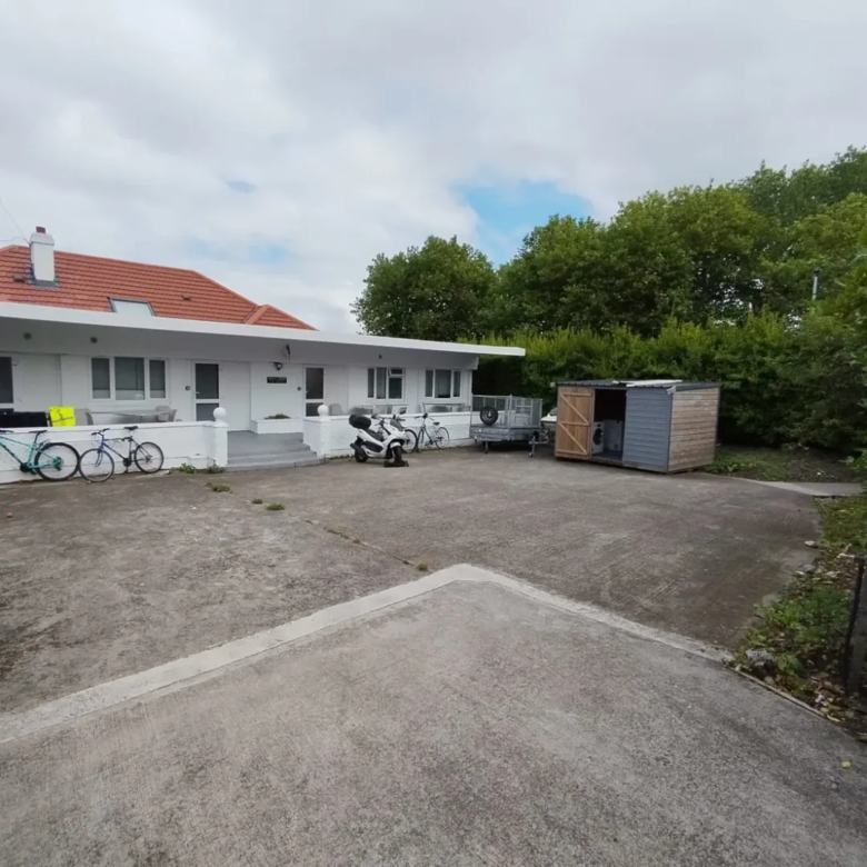 White bungalow with red roof, bikes, scooter, trailer, shed, and concrete driveway on an overcast day.